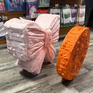 K&ouml;nnte beinhalten: Eine rosa Schleifen-Pi&ntilde;ata und eine orangefarbene Kreis-Pi&ntilde;ata. Die Schleifen-Pi&ntilde;ata besteht aus geschichtetem rosa Papier und hat eine gro&szlig;e Schleife in der Mitte. Die orangefarbene Pi&ntilde;ata besteht aus orangefarbenen Papierfransen.