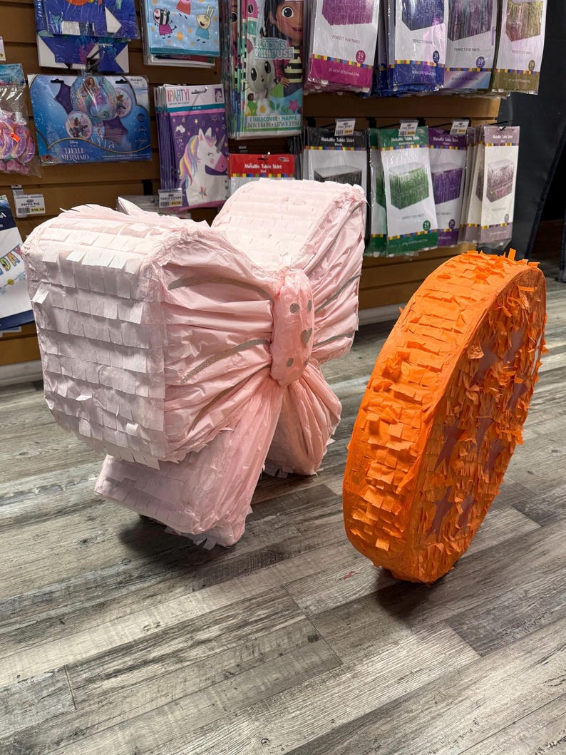 K&ouml;nnte beinhalten: Eine rosa Schleifen-Pi&ntilde;ata und eine orangefarbene Kreis-Pi&ntilde;ata. Die Schleifen-Pi&ntilde;ata besteht aus geschichtetem rosa Papier und hat eine gro&szlig;e Schleife in der Mitte. Die orangefarbene Pi&ntilde;ata besteht aus orangefarbenen Papierfransen.