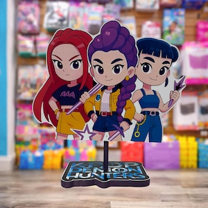 May include: A colorful K-Pop Demon Hunters stand-up figure featuring three cartoon characters with different hair colors and outfits. The base of the figure has the text "K-Pop Demon Hunters" in blue and white. The characters are holding weapons.