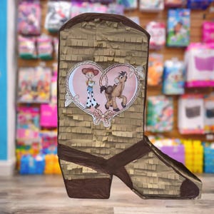 May include: A brown boot-shaped pi&ntilde;ata with a decorative heart featuring cartoon characters. The pi&ntilde;ata is made of brown paper with a fringed texture and dark brown accents. The heart has a pink background and white ribbon details.