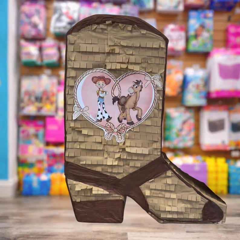 May include: A brown boot-shaped pi&ntilde;ata with a decorative heart featuring cartoon characters. The pi&ntilde;ata is made of brown paper with a fringed texture and dark brown accents. The heart has a pink background and white ribbon details.