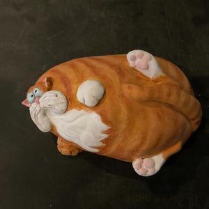 May include: A ceramic ornament of a ginger tabby cat lying on its back. The cat has white patches on its belly and paws, with blue eyes and a surprised expression. The cat's fur is striped with darker orange lines.