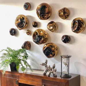 May include: A collection of twelve irregular, metallic bronze-colored wall sculptures with a reflective, liquid-like surface. Below, a silver balloon dog figurine and a black-framed hourglass sit on a dark wooden cabinet next to a green fern.