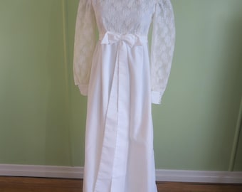 Puede incluir: Un vestido de novia blanco con corpiño de encaje y mangas largas de encaje. El vestido presenta un lazo en la cintura y una falda larga y fluida. Un tocado floral adorna la cabeza del maniquí. El vestido se exhibe en un maniquí.