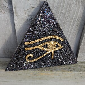 May include: A triangular, black decorative piece with a textured surface and a gold-coloured Eye of Horus symbol. The surface has iridescent flecks of blue, purple, and silver. The Eye of Horus is a symbol of protection and good health.
