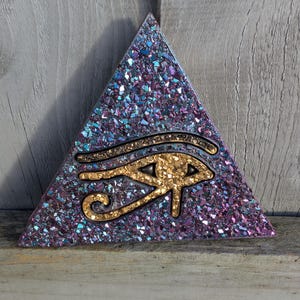 May include: A triangular decorative piece with a glittery purple and blue surface. A gold-coloured Eye of Horus symbol is centered on the triangle. The piece is displayed on a wooden surface.