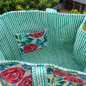 Op de afbeelding: Een handgemaakte tote bag met een groen-witte gestreepte buitenkant en een bloemeninterieur. De tas heeft een klein vakje met een bloemenpatroon van rode rozen en blauwe bladeren. De handvatten zijn ook gestreept.