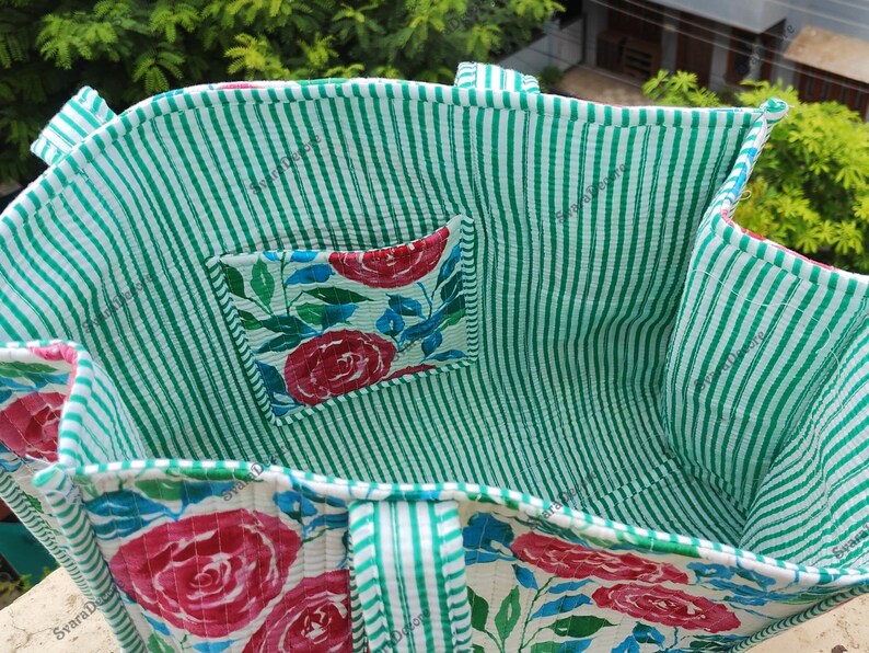 Op de afbeelding: Een handgemaakte tote bag met een groen-witte gestreepte buitenkant en een bloemeninterieur. De tas heeft een klein vakje met een bloemenpatroon van rode rozen en blauwe bladeren. De handvatten zijn ook gestreept.