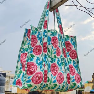 Op de afbeelding: Een grote draagtas met een bloemenpatroon van rode rozen en blauwe bladeren op een witte achtergrond. De tas heeft groene gestreepte randen en handvatten. De tas hangt buiten tegen een bewolkte lucht.