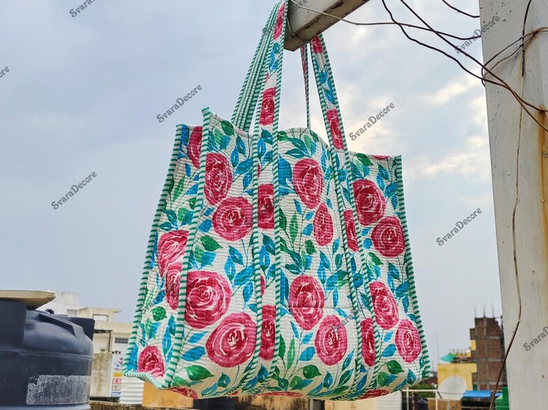 Op de afbeelding: Een grote draagtas met een bloemenpatroon van rode rozen en blauwe bladeren op een witte achtergrond. De tas heeft groene gestreepte randen en handvatten. De tas hangt buiten tegen een bewolkte lucht.