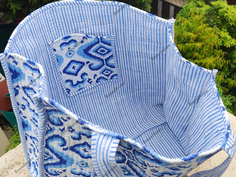 K&ouml;nnte beinhalten: Offene Tragetasche mit blauem und wei&szlig;em Muster. Die Tasche hat eine Tasche mit geometrischem Muster und einen gestreiften Innenraum. Die Griffe und der Besatz sind ebenfalls gemustert. Die Tasche ist aus weichem, gestepptem Stoff gefertigt.