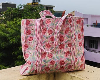 Könnte beinhalten: Eine große, gesteppte Tragetasche mit einem Blumenmuster in Rosa- und Rottönen. Die Tasche hat eine rechteckige Form mit stabilen Griffen und einer passenden rosa-weiß gestreiften Bordüre. Die Tasche ist aus einem weichen, gesteppten Stoff gefertigt.