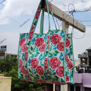 Op de afbeelding: Een grote, rechthoekige draagtas met een bloemenpatroon van rode rozen en blauwe bladeren op een witte achtergrond. De tas heeft bijpassende handvatten en een rand in een groen-wit gestreept patroon. De tas hangt buiten tegen een bewolkte lucht. Afmetingen: 40 cm x 40 cm.