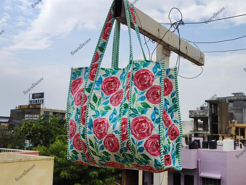 Op de afbeelding: Een grote, rechthoekige draagtas met een bloemenpatroon van rode rozen en blauwe bladeren op een witte achtergrond. De tas heeft bijpassende handvatten en een rand in een groen-wit gestreept patroon. De tas hangt buiten tegen een bewolkte lucht. Afmetingen: 40 cm x 40 cm.