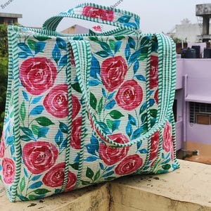 Op de afbeelding: Een bloemen draagtas met een patroon van rode rozen, blauwe bladeren en groene accenten op een witte achtergrond. De tas heeft groene gestreepte randen en handvatten. Ideaal voor het dragen van spullen.