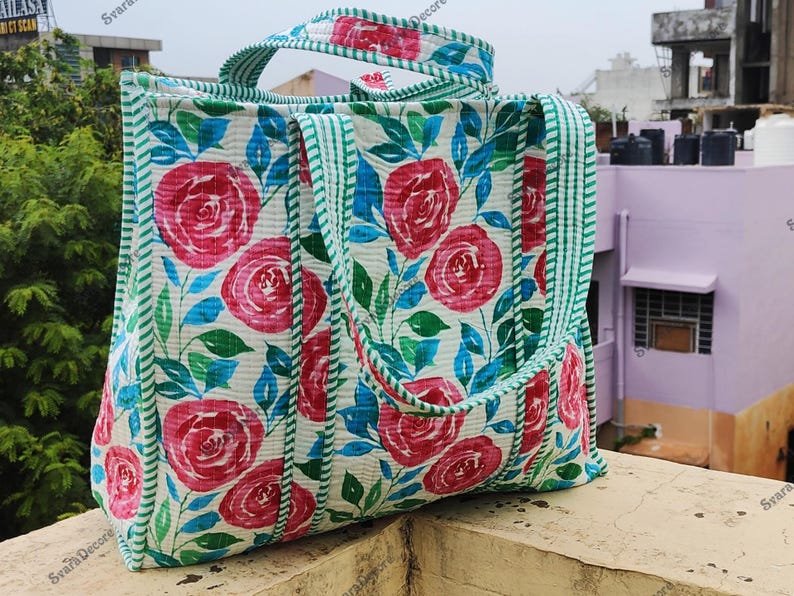 Op de afbeelding: Een bloemen draagtas met een patroon van rode rozen, blauwe bladeren en groene accenten op een witte achtergrond. De tas heeft groene gestreepte randen en handvatten. Ideaal voor het dragen van spullen.