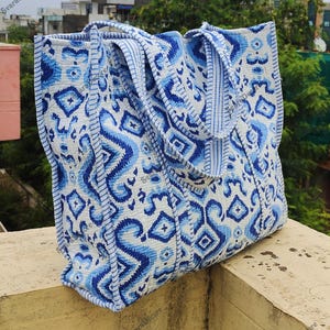 K&ouml;nnte beinhalten: Eine gro&szlig;e Tragetasche mit einem wei&szlig;en und blauen Muster. Die Tasche hat ein sich wiederholendes Muster aus geometrischen Formen und Wirbeln in verschiedenen Blaut&ouml;nen. Die Tasche hat zwei Griffe und ist aus einem robusten Stoff gefertigt.