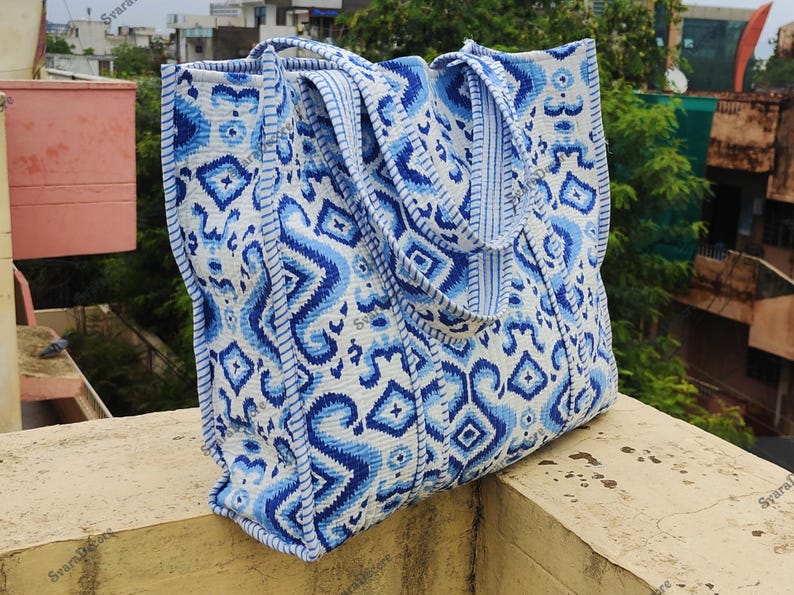 K&ouml;nnte beinhalten: Eine gro&szlig;e Tragetasche mit einem wei&szlig;en und blauen Muster. Die Tasche hat ein sich wiederholendes Muster aus geometrischen Formen und Wirbeln in verschiedenen Blaut&ouml;nen. Die Tasche hat zwei Griffe und ist aus einem robusten Stoff gefertigt.