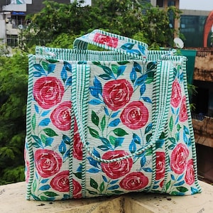 Op de afbeelding: Een rechthoekige draagtas met een bloemenpatroon. De tas heeft grote rode rozen, blauwe bladeren en groene accenten op een witte achtergrond. De handvatten en randen zijn afgewerkt met groen-witte strepen.