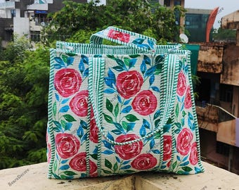 Könnte beinhalten: Eine rechteckige Tragetasche mit floralem Muster. Die Tasche zeigt große rote Rosen, blaue Blätter und grüne Akzente auf weißem Grund. Die Griffe und Kanten sind mit grün-weißen Streifen verziert.