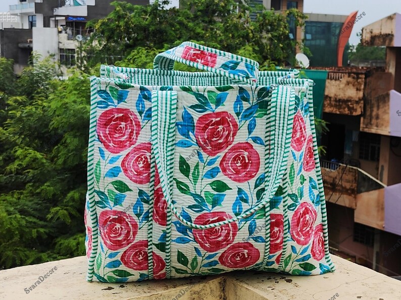 Op de afbeelding: Een rechthoekige draagtas met een bloemenpatroon. De tas heeft grote rode rozen, blauwe bladeren en groene accenten op een witte achtergrond. De handvatten en randen zijn afgewerkt met groen-witte strepen.