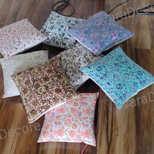 May include: Assortment of eight decorative throw pillows with various floral patterns in shades of pink, blue, green, and brown. The pillows are arranged on a wooden floor.