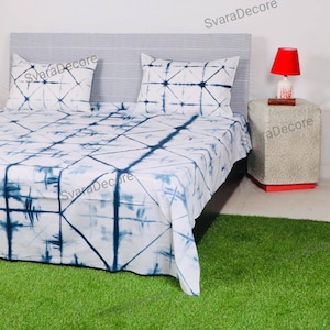May include: A white bed with a blue and white tie-dye patterned bedspread and two matching pillowcases. The bed is made up with a gray and white striped headboard. A small side table with a red lamp sits next to the bed.