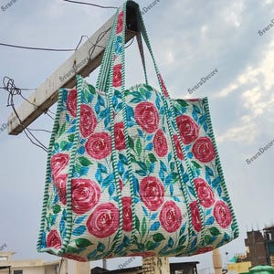 Op de afbeelding: Een grote, gewatteerde draagtas met een bloemenrozenpatroon in roze, blauw en groen op een witte achtergrond. De tas heeft bijpassende handvatten en afwerking. De tas hangt buiten tegen een bewolkte lucht.