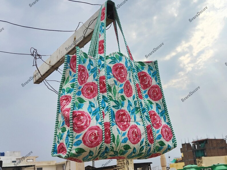 Op de afbeelding: Een grote, gewatteerde draagtas met een bloemenrozenpatroon in roze, blauw en groen op een witte achtergrond. De tas heeft bijpassende handvatten en afwerking. De tas hangt buiten tegen een bewolkte lucht.