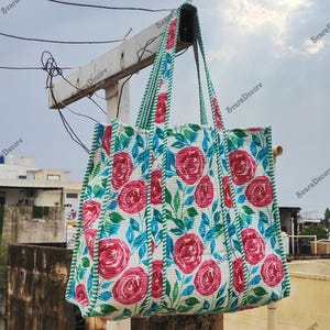 Op de afbeelding: Een grote draagtas met een bloemenrozenpatroon in rood, wit en blauw. De tas heeft groen gestreepte handvatten en randen. De tas is gemaakt van gewatteerde stof en hangt buiten.