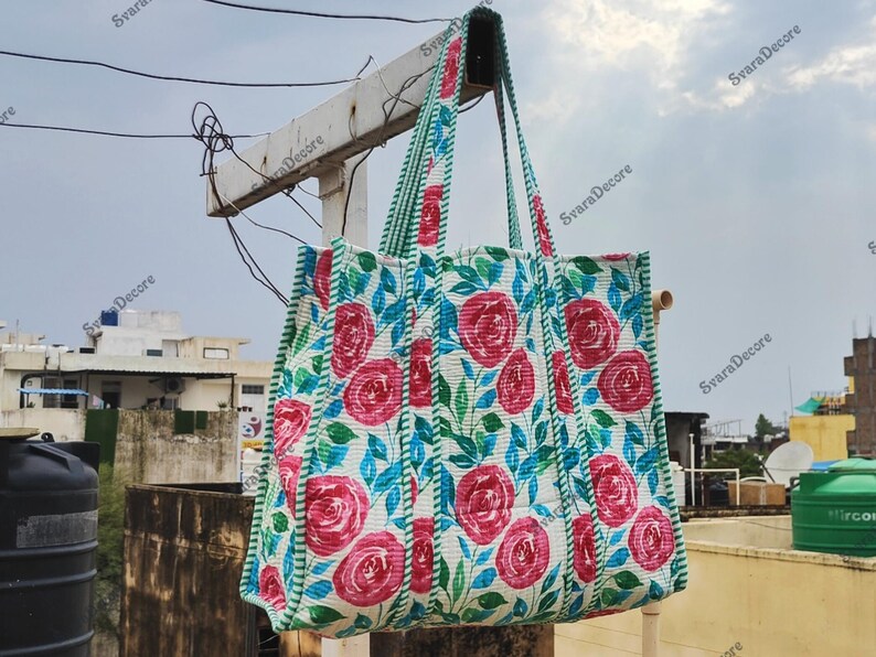 Op de afbeelding: Een grote draagtas met een bloemenrozenpatroon in rood, wit en blauw. De tas heeft groen gestreepte handvatten en randen. De tas is gemaakt van gewatteerde stof en hangt buiten.