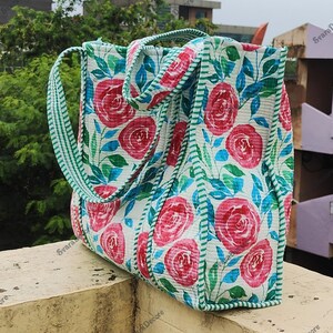 Op de afbeelding: Een bloemen draagtas met grote roze rozen op een witte achtergrond. De tas heeft groene en blauwe bladeren, een groen gestreepte rand en groen gestreepte handvatten. De tas is gemaakt van een gewatteerde stof.