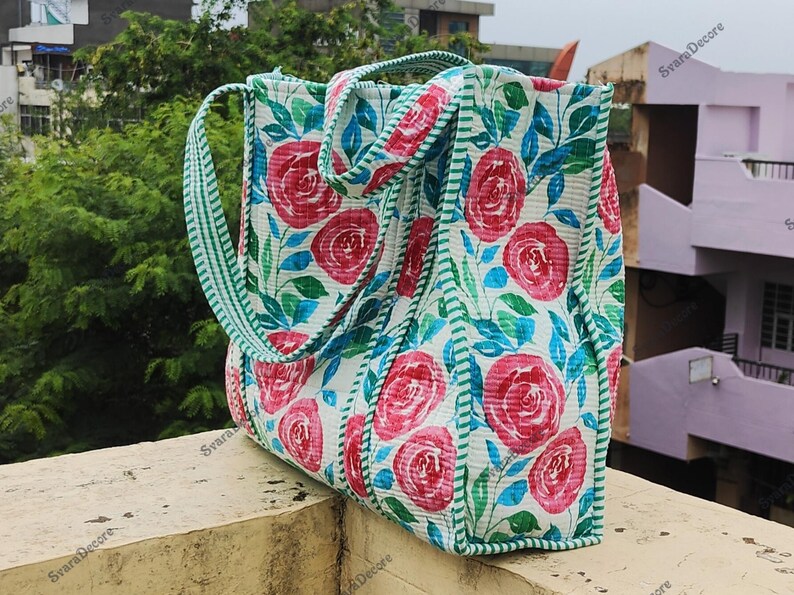 Op de afbeelding: Een bloemen draagtas met grote roze rozen op een witte achtergrond. De tas heeft groene en blauwe bladeren, een groen gestreepte rand en groen gestreepte handvatten. De tas is gemaakt van een gewatteerde stof.
