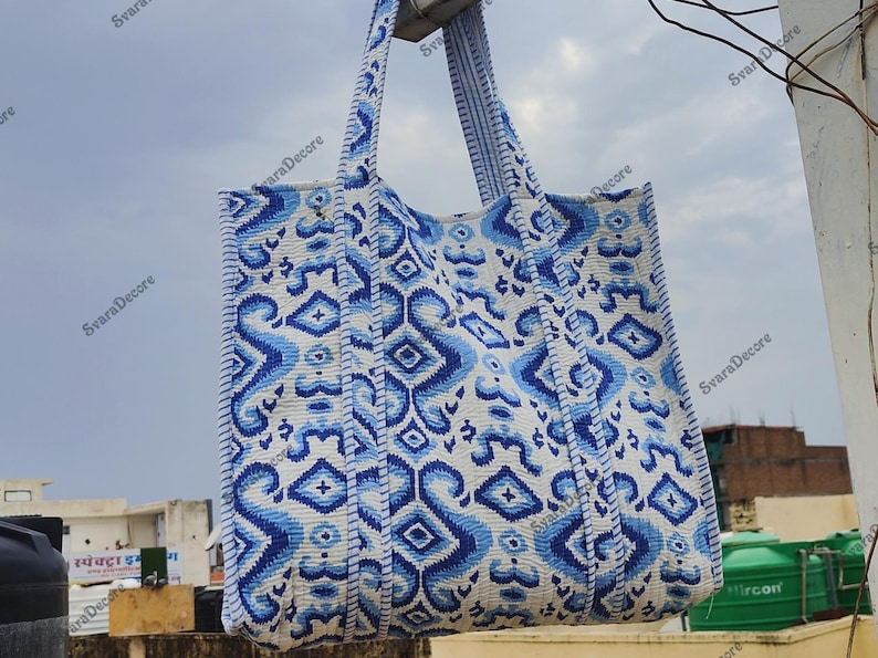 K&ouml;nnte beinhalten: Eine gro&szlig;e Tragetasche mit einem blau-wei&szlig;en geometrischen Muster. Die Tasche hat zwei Griffe und ist aus einem strukturierten Stoff gefertigt. Das Design zeigt sich wiederholende Formen und Linien.