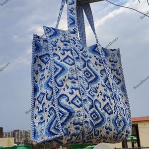 K&ouml;nnte beinhalten: Eine gro&szlig;e Tragetasche mit einem blau-wei&szlig;en geometrischen Muster. Die Tasche hat zwei Griffe und ist aus einem gesteppten Stoff gefertigt. Das Design zeigt Diamanten und abstrakte Formen. Die Tasche h&auml;ngt drau&szlig;en vor einem bew&ouml;lkten Himmel.