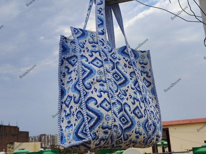K&ouml;nnte beinhalten: Eine gro&szlig;e Tragetasche mit einem blau-wei&szlig;en geometrischen Muster. Die Tasche hat zwei Griffe und ist aus einem gesteppten Stoff gefertigt. Das Design zeigt Diamanten und abstrakte Formen. Die Tasche h&auml;ngt drau&szlig;en vor einem bew&ouml;lkten Himmel.