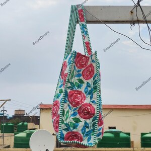 Op de afbeelding: Een draagtas met bloemenpatroon met grote rode rozen, groene bladeren en blauwe accenten op een witte achtergrond. De tas heeft groen-wit gestreepte handvatten en randen. De tas hangt buiten.