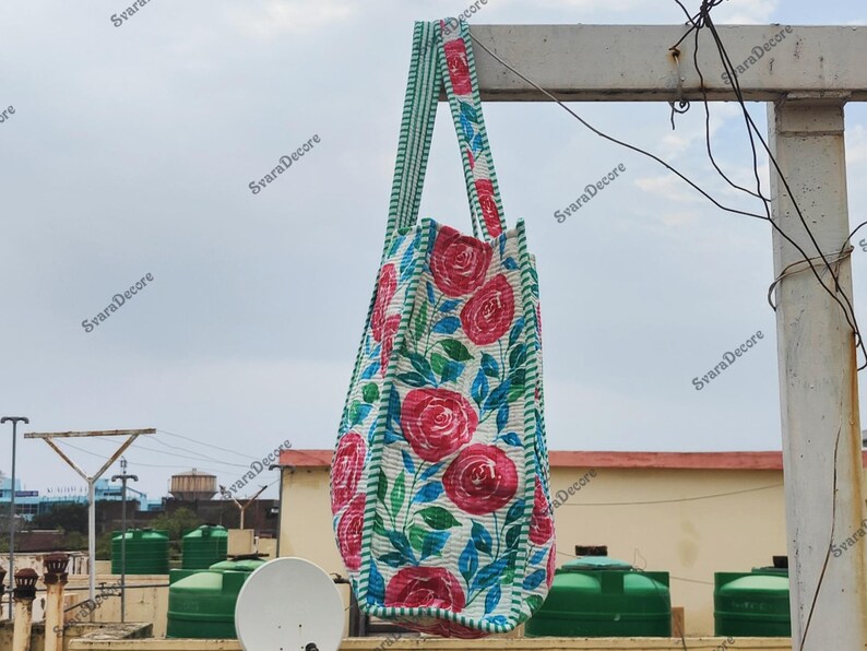 Op de afbeelding: Een draagtas met bloemenpatroon met grote rode rozen, groene bladeren en blauwe accenten op een witte achtergrond. De tas heeft groen-wit gestreepte handvatten en randen. De tas hangt buiten.
