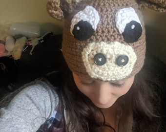 May include: A brown crocheted cow hat with white accents and black button eyes. The hat is worn by a person with long brown hair.