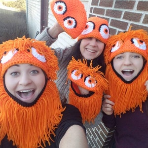 May include: Three people wearing orange and black crocheted hats with fuzzy orange hair and large white eyes. The hats have a black crocheted chin strap.