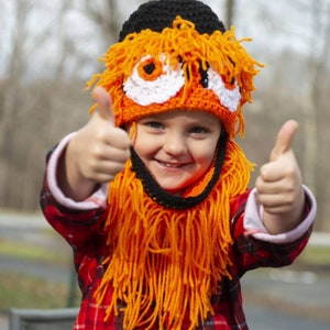 May include: A child wearing a bright orange and black crocheted hat with a long, shaggy orange fringe. The hat has white and orange eyes and a black nose. The child is giving a thumbs up with both hands.