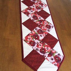 Puede incluir: Un camino de mesa acolchado con un patrón de diamantes burdeos y blancos. El camino de mesa presenta un estampado floral con flores rojas, rosas y moradas.