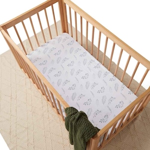 Pode incluir: Um berço de madeira clara com um lençol branco com um padrão de folhas cinzentas. Uma manta verde escura está sobre o lado do berço. O berço está sobre um tapete creme.
