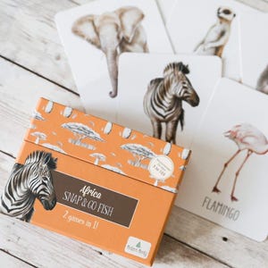 Könnte beinhalten: Ein orangefarbener Karton mit der Aufschrift "Africa SNAP & GO FISH" und "2 Spiele in 1!" ist von Tierkarten umgeben. Die Karten zeigen Illustrationen eines Zebras, eines Elefanten, eines Flamingos und eines Erdmännchens. Der Karton ist mit einem Savannenmuster verziert.