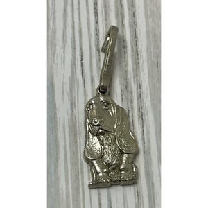 May include: A silver basset hound charm with a loop for attachment. The charm shows a seated dog with long ears and a detailed face. The charm is made of a shiny metal.