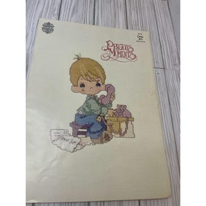 May include: Cross-stitch pattern featuring a cartoon boy with blonde hair, holding a pink telephone. The design includes the words "Precious Moments" and "Dear Jon". The boy is wearing a green shirt and blue jeans. The background is a light cream color.