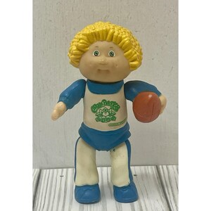 Op de afbeelding: Een kleine plastic pop met geel gevormd haar en groene ogen. De pop draagt een wit shirt met een groen Cabbage Patch Kids-logo en blauwe tekst, een blauwe broek en een witte broek met blauwe strepen. Het houdt een bruine football in de linkerhand.