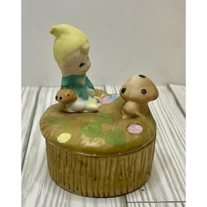 Puede incluir: Una caja de baratijas de cerámica con un diseño caprichoso. La caja tiene forma de hongo con una figura de gnomo sentado y un acento de hongo. El gnomo lleva un sombrero amarillo y una chaqueta verde. La caja tiene un exterior marrón y texturizado.