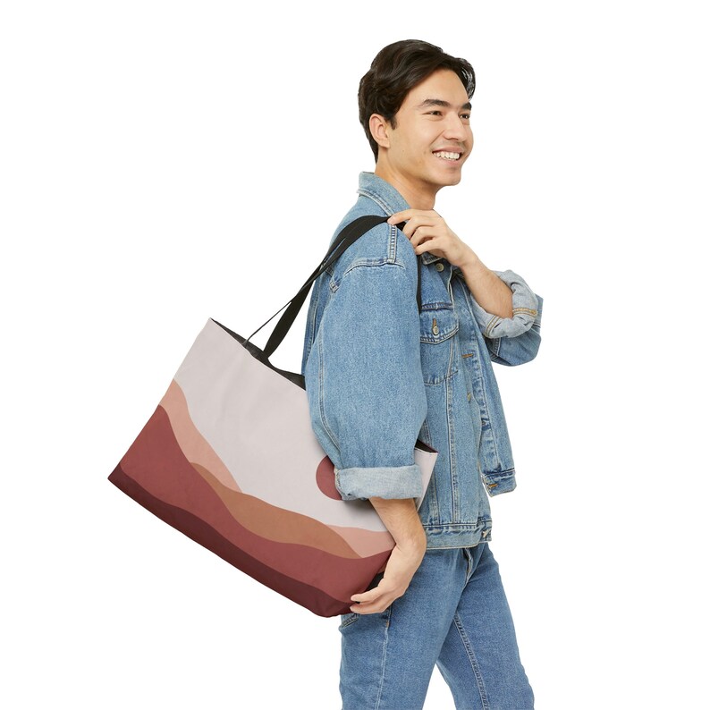 May include: A large tote bag with a landscape design in shades of red, brown, and beige. The bag has black straps and a black interior. The design features a sun and layered hills. The bag is carried over the shoulder.