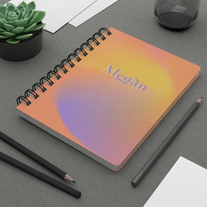May include: A spiral bound notebook with a peach and purple ombre cover. The notebook has the name "Megan" written in a stylized font on the front.
